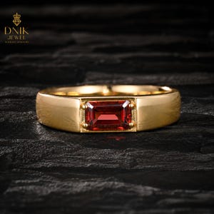 May include: A gold ring with a rectangular, deep red gemstone at the center. The ring has a simple, elegant design with a polished finish. The gemstone is securely set within the ring's band, showcasing a classic jewelry style.