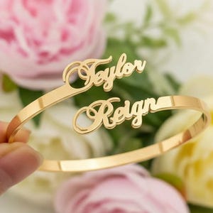 Puede incluir: Una pulsera de puño dorada con los nombres "Taylor" y "Reign" en cursiva. La pulsera se muestra frente a un fondo borroso de flores rosas y amarillas.