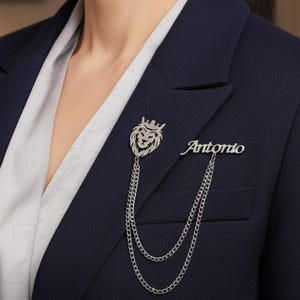 Puede incluir: Broche plateado con la cabeza de un león coronado y el nombre "Antonio". El broche está sujeto a una doble cadena, exhibido en una blazer azul marino sobre una camisa blanca. Un accesorio elegante para chaquetas y blazers.