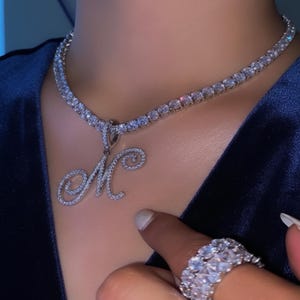 Puede incluir: Un collar de tenis de diamantes plateado con un colgante con la letra "M". El colgante y el collar están adornados con pequeñas piedras preciosas transparentes. También se ve un anillo de diamantes a juego.