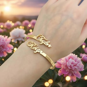May include: Gold-coloured cuff bracelet with the names "Taylor" and "Reign" in a cursive font. The bracelet is displayed on a wrist, with a blurred floral background featuring pink and white flowers.