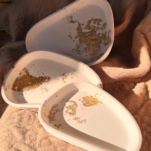 May include: Set of three white, oval-shaped trays with gold leaf accents. The trays are arranged in a layered composition, showcasing their varying sizes and the decorative gold leaf patterns. The trays are on a peach-colored surface.