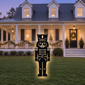 May include: A black silhouette of a nutcracker figure with a glowing yellow outline, featuring a tall hat, a mustache, and a uniform with horizontal lines, standing upright on a green lawn in front of a white house with a porch.