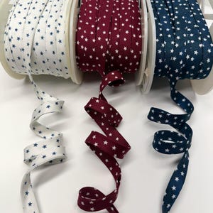 May include: Three spools of star-patterned bias tape in white with navy stars, burgundy with white stars, and navy with white stars. The tape is unrolled in a wavy pattern, showcasing the star design.