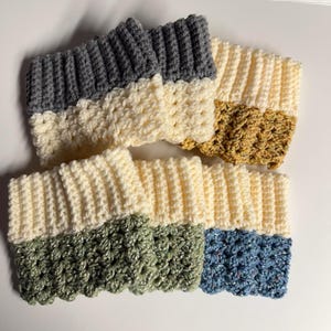 May include: A collection of six crocheted wrist warmers in various colors. The wrist warmers are two-toned, with a ribbed top section and a textured bottom section. Colors include gray, cream, olive green, blue, and gold.