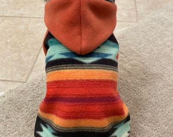 May include: A small white dog wearing a hooded fleece sweater with a colourful southwestern pattern. The sweater has a brown leather tag that says "Hand Made".