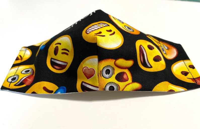 May include: Black face mask with a yellow emoji pattern. The emojis include winking, smiling, and heart-eyed faces.