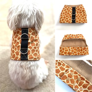 Op de afbeelding: Een bruin en tan hondenharnas met een giraffenprint. Het harnas heeft een zwarte band met twee zilveren D-ringen. Het harnas wordt getoond op een kleine witte hond.