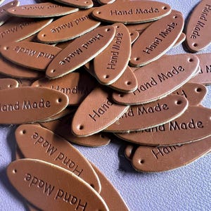 Op de afbeelding: Een stapel ovale, bruine leren labels met de tekst "Hand Made" in reliëf. Elk label heeft twee kleine gaatjes aan de uiteinden. De labels liggen verspreid op een lichtpaarse ondergrond.