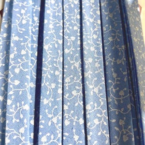 Puede incluir: Primer plano de una tela azul claro con un estampado floral blanco. La tela está plisada, creando líneas verticales. Una cinta azul más oscura es visible en el lado derecho. La tela probablemente se utiliza para ropa o decoración del hogar.