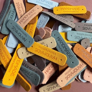 May include: A collection of rectangular faux leather tags in various colors including yellow, blue, brown, and tan. Each tag features the embossed text "HANDMADE Limited edition" and has a small hole punched at each end for attachment purposes.