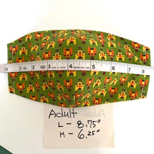 May include: Green fabric face mask with a repeating pattern of orange and yellow cartoon cats. The mask is measured with a tape measure and the measurements are written on a white piece of paper: Adult, L - 8.75", H - 6.25".
