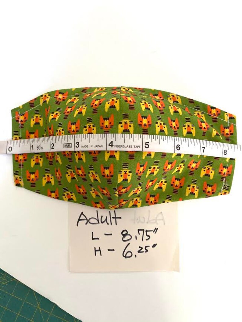 May include: Green fabric face mask with a repeating pattern of orange and yellow cartoon cats. The mask is measured with a tape measure and the measurements are written on a white piece of paper: Adult, L - 8.75", H - 6.25".