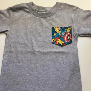 Puede incluir: Camiseta gris de manga larga con un bolsillo que presenta una imagen de c&oacute;mic de Capit&aacute;n Am&eacute;rica en una escena de lucha. El bolsillo est&aacute; cosido en la parte delantera de la camiseta.