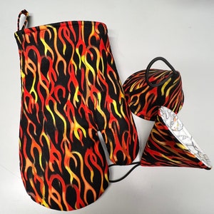 May include: A set of three oven mitts and pot holders with a flame pattern in red, yellow, and black. The mitts have a black background with yellow and red flames. The pot holders have a black background with yellow and red flames and a white lining.