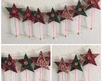 May include: A festive garland of red and green fabric stars with gold bells hanging from each star. The stars are sewn together with white twine and the bells are attached with red and white twine.