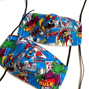 May include: Two blue fabric face masks with black elastic ear loops. The masks feature a comic book pattern with images of superheroes like Spider-Man, Captain America, and The Hulk. The text on the masks includes "The Incredible Hulk", "Captain America", and "Amazing Spider-Man".