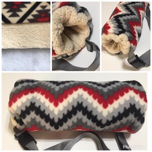 May include: A fleece hand warmer with a gray, red, black, and white chevron pattern. The interior is a cream-colored sherpa material. It has a gray adjustable strap with a black buckle.