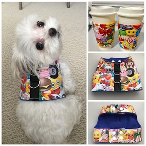May include: A white dog wearing a blue and black harness with a colorful emoji pattern. The harness has two silver rings and a black strap. The harness features a variety of emojis, including a smiley face, a hamburger, a car, and a heart. The dog is looking up at the camera.