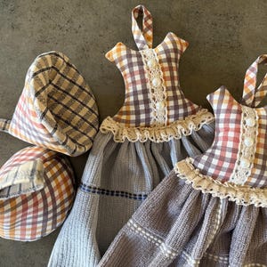 Könnte beinhalten: Eine Sammlung von Küchenaccessoires, darunter zwei handtuchförmige Kleider zum Aufhängen und zwei passende Topflappen. Die Handtücher und Topflappen weisen ein Karomuster in Braun-, Orange- und Weißtönen mit Spitzen- und Knopfd details auf.
