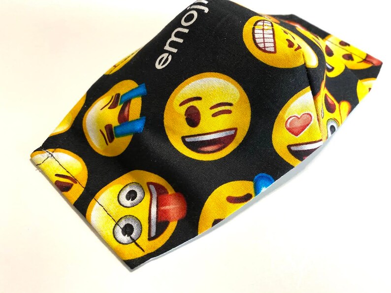 May include: Black fabric face mask with a repeating pattern of yellow emoji faces. The text "emoji" is printed in white on the top of the mask.