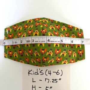 May include: Green fabric face mask with a repeating pattern of orange and yellow cartoon cats. The mask is measured with a tape measure and the measurements are written on a white card: Kid's (4-6) L - 7.25" H - 5"