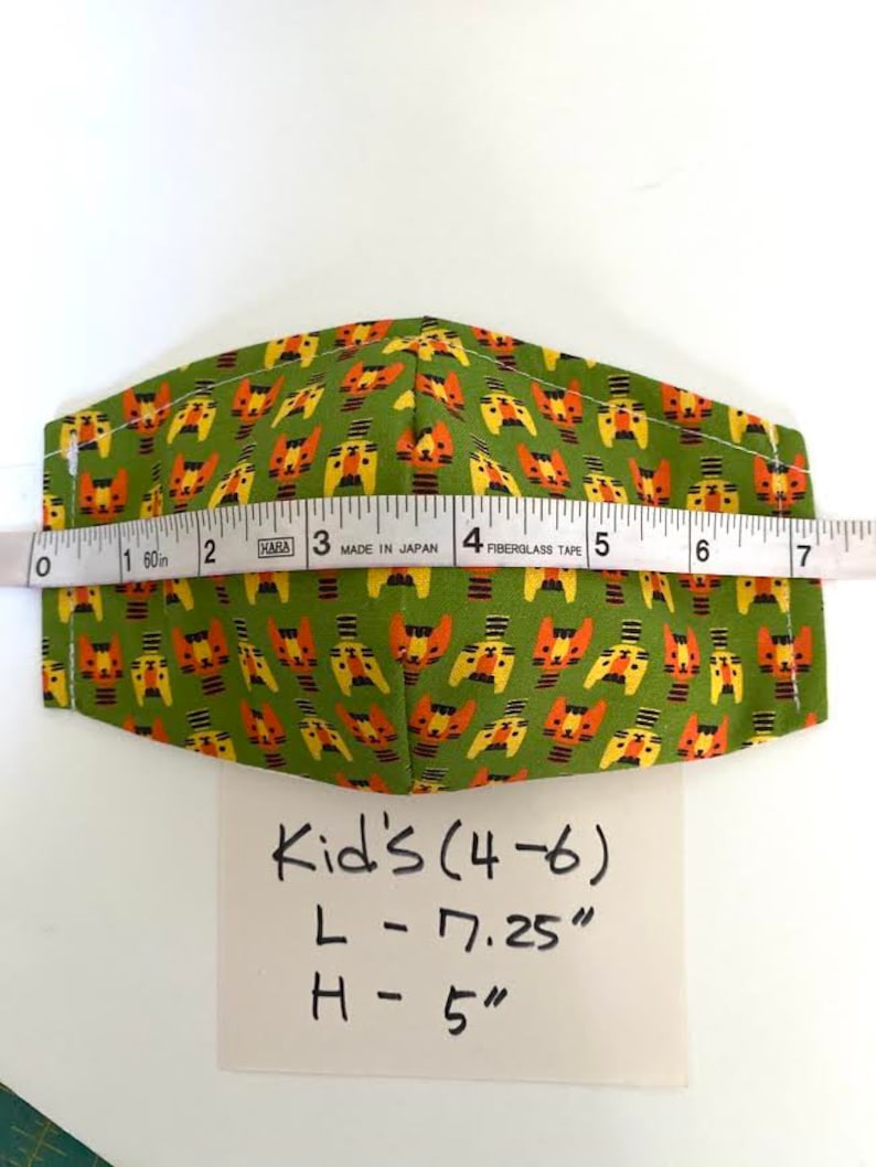 May include: Green fabric face mask with a repeating pattern of orange and yellow cartoon cats. The mask is measured with a tape measure and the measurements are written on a white card: Kid's (4-6) L - 7.25" H - 5"