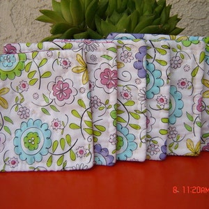 May include: Set of eight white fabric coasters with a floral pattern in shades of pink, blue, green, and yellow. The coasters are lined with a green trim.