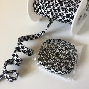 May include: A spool of black and white houndstooth patterned elastic ribbon. The ribbon is wound around a white spool and is also shown in a separate plastic bag.
