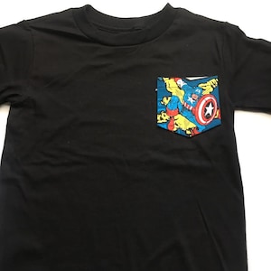 Puede incluir: Camiseta negra con un bolsillo que presenta una imagen de c&oacute;mic de Capit&aacute;n Am&eacute;rica con un traje azul, rojo y amarillo.