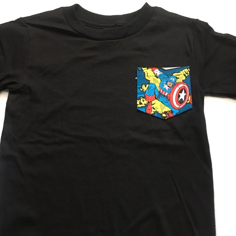 Puede incluir: Camiseta negra con un bolsillo que presenta una imagen de c&oacute;mic de Capit&aacute;n Am&eacute;rica con un traje azul, rojo y amarillo.