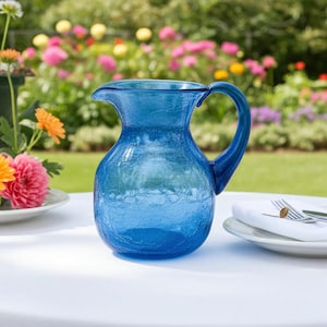 May include: A blue glass pitcher with a crackle finish sits on a white tablecloth. The pitcher has a curved handle and a spout for pouring. The background features a garden with colorful flowers.