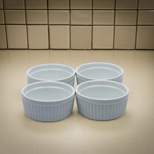 May include: A set of four light blue ceramic ramekins arranged in a square formation. Each round dish features a smooth interior and a vertically ribbed exterior texture, providing a consistent look for these small, open-top baking containers.