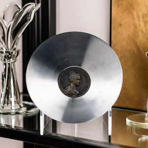 May include: A circular, brushed silver-toned metal plate featuring a centered, dark bronze-colored medallion. The medallion displays a relief profile of a person with an ornate hairstyle, facing right, set against a dark, textured circular background.