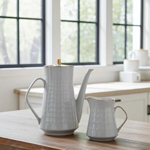 May include: A tall, light gray porcelain coffee pot and a matching creamer pitcher. Both pieces feature a subtle, repeating geometric embossed pattern on the exterior, gold-toned rims, and a gold-toned knob on the coffee pot lid.