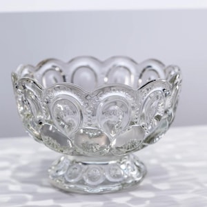 May include: A clear glass bowl featuring a repeating moon and star pattern with a scalloped rim. The bowl sits on a circular pedestal base and is constructed from pressed glass with a textured surface that catches light across its rounded exterior.