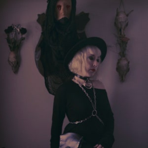 May include: A woman wearing a black hat and black clothing with a silver chain and a fringe bag. She is sitting on a fur rug in front of a deer head with red eyes.