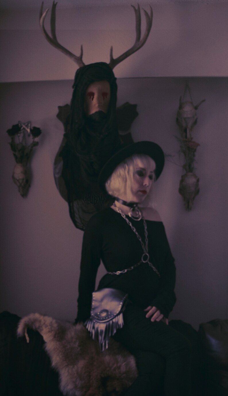 May include: A woman wearing a black hat and black clothing with a silver chain and a fringe bag. She is sitting on a fur rug in front of a deer head with red eyes.