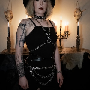May include: A woman wearing a black hat, black tank top, and a silver chain harness. She has a tattoo on her arm and is standing in front of a dark background with candles and skulls.
