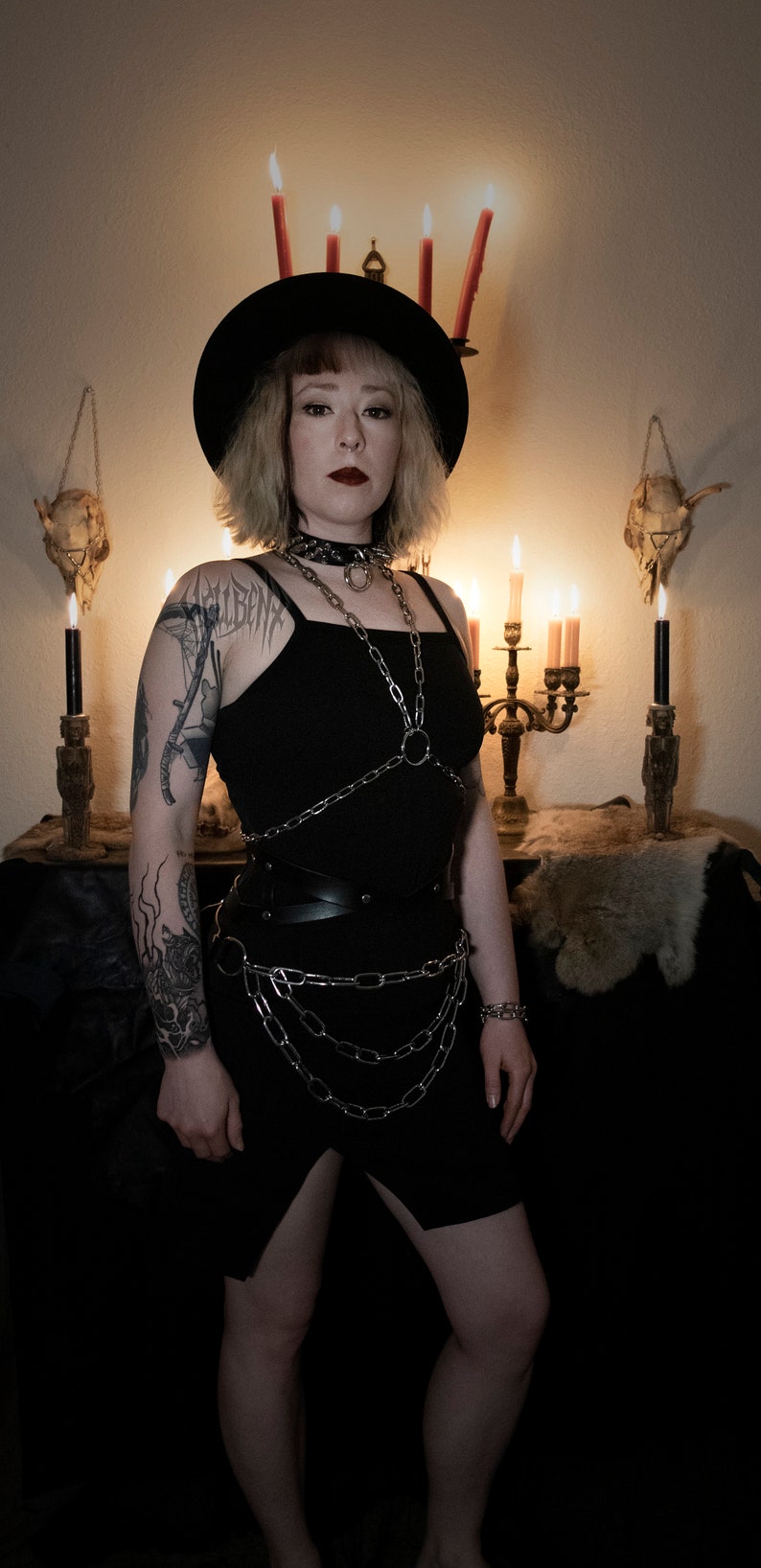 May include: A woman wearing a black hat, black tank top, and a silver chain harness. She has a tattoo on her arm and is standing in front of a dark background with candles and skulls.
