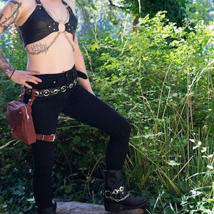 May include: A woman wearing a black leather halter top with silver hardware, black jeans, a brown leather belt with silver buckles, and black boots with silver buckles. She is standing in a forest setting.