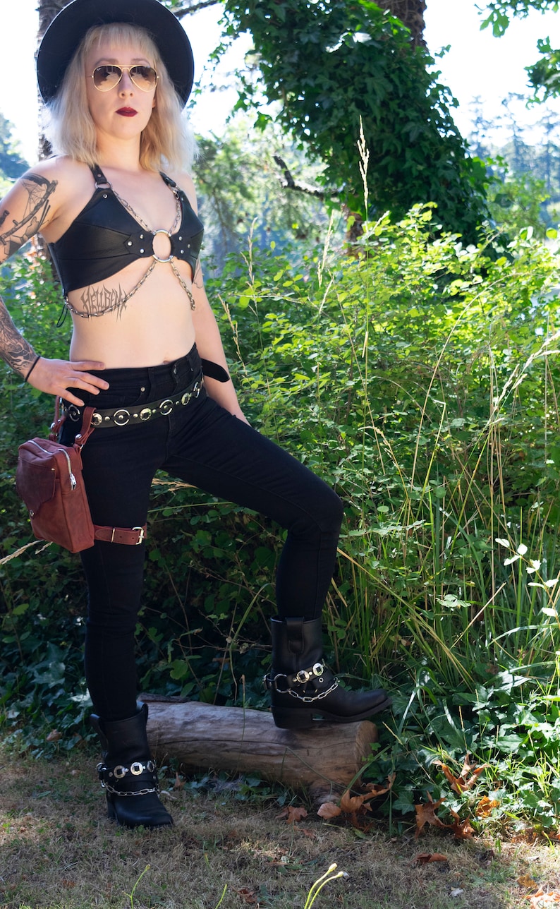 May include: A woman wearing a black leather halter top with silver hardware, black jeans, a brown leather belt with silver buckles, and black boots with silver buckles. She is standing in a forest setting.