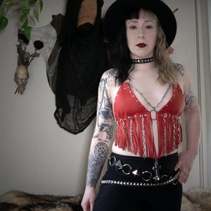 Puede incluir: Una mujer con un top rojo de cuero con flecos y una cadena plateada, y un sombrero negro. Tambi&eacute;n lleva un collar negro de gargantilla y un cintur&oacute;n de tachuelas plateadas.