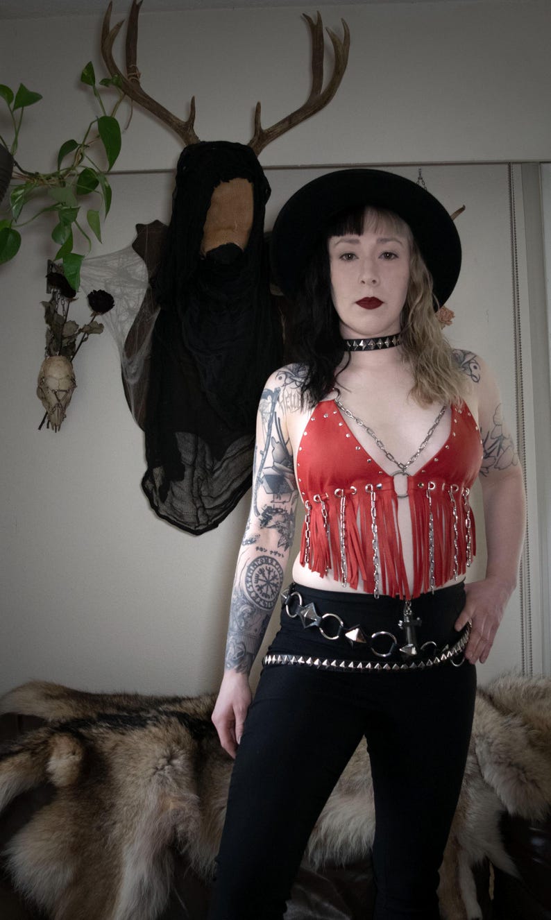Puede incluir: Una mujer con un top rojo de cuero con flecos y una cadena plateada, y un sombrero negro. Tambi&eacute;n lleva un collar negro de gargantilla y un cintur&oacute;n de tachuelas plateadas.