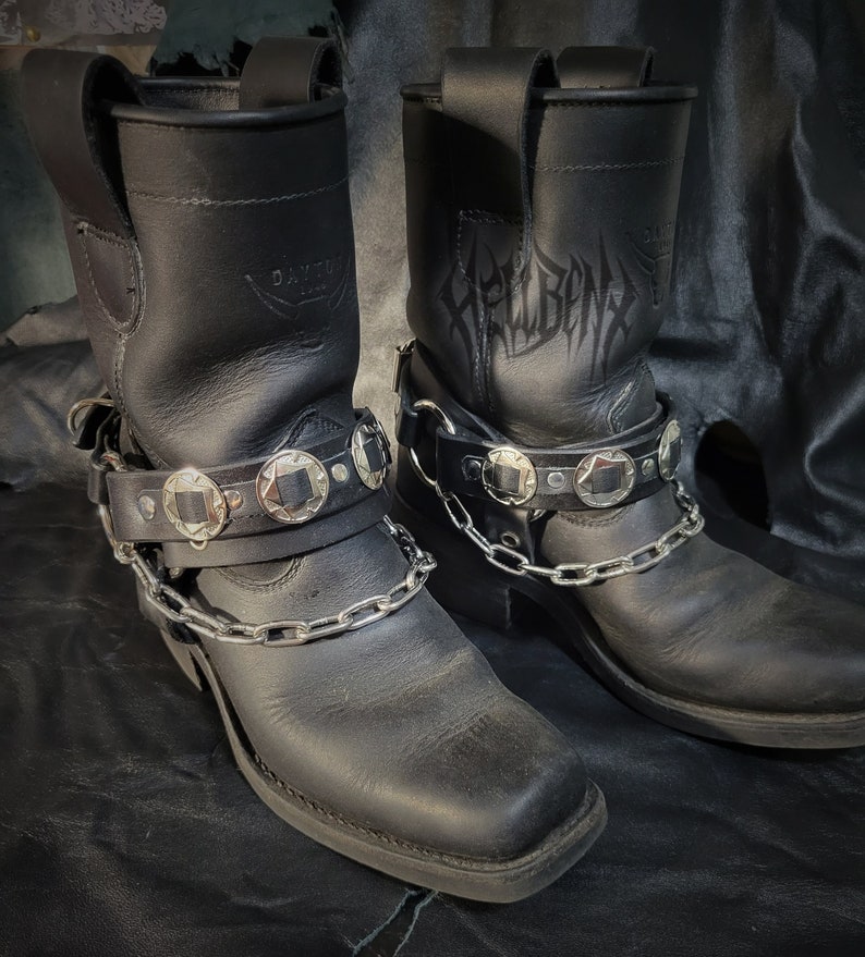 May include: A pair of black leather motorcycle boots with silver buckles and chains. The boots have a pointed toe and a chunky heel. The boots are decorated with the word "Hellbent" on the side.