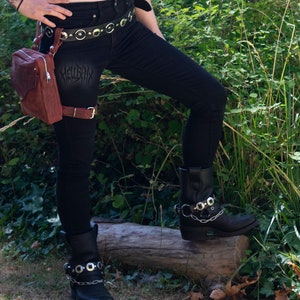 May include: A person wearing black jeans with a brown leather belt and a brown leather thigh holster with a small brown bag attached. The person is wearing black boots with silver buckles and chains.
