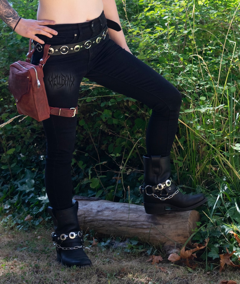 May include: A person wearing black jeans with a brown leather belt and a brown leather thigh holster with a small brown bag attached. The person is wearing black boots with silver buckles and chains.