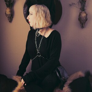 May include: A woman wearing a black top with a silver chain and a black leather bag sits on a furry rug. She is wearing black boots and has short blonde hair. A taxidermied deer head with red eyes is mounted on the wall behind her.