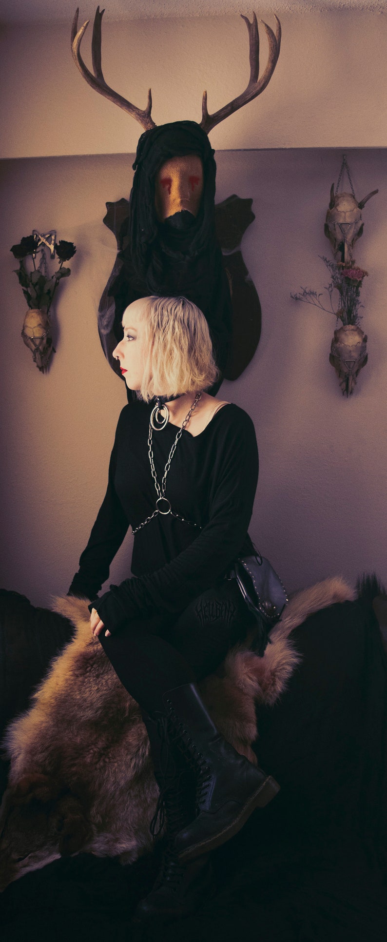 May include: A woman wearing a black top with a silver chain and a black leather bag sits on a furry rug. She is wearing black boots and has short blonde hair. A taxidermied deer head with red eyes is mounted on the wall behind her.