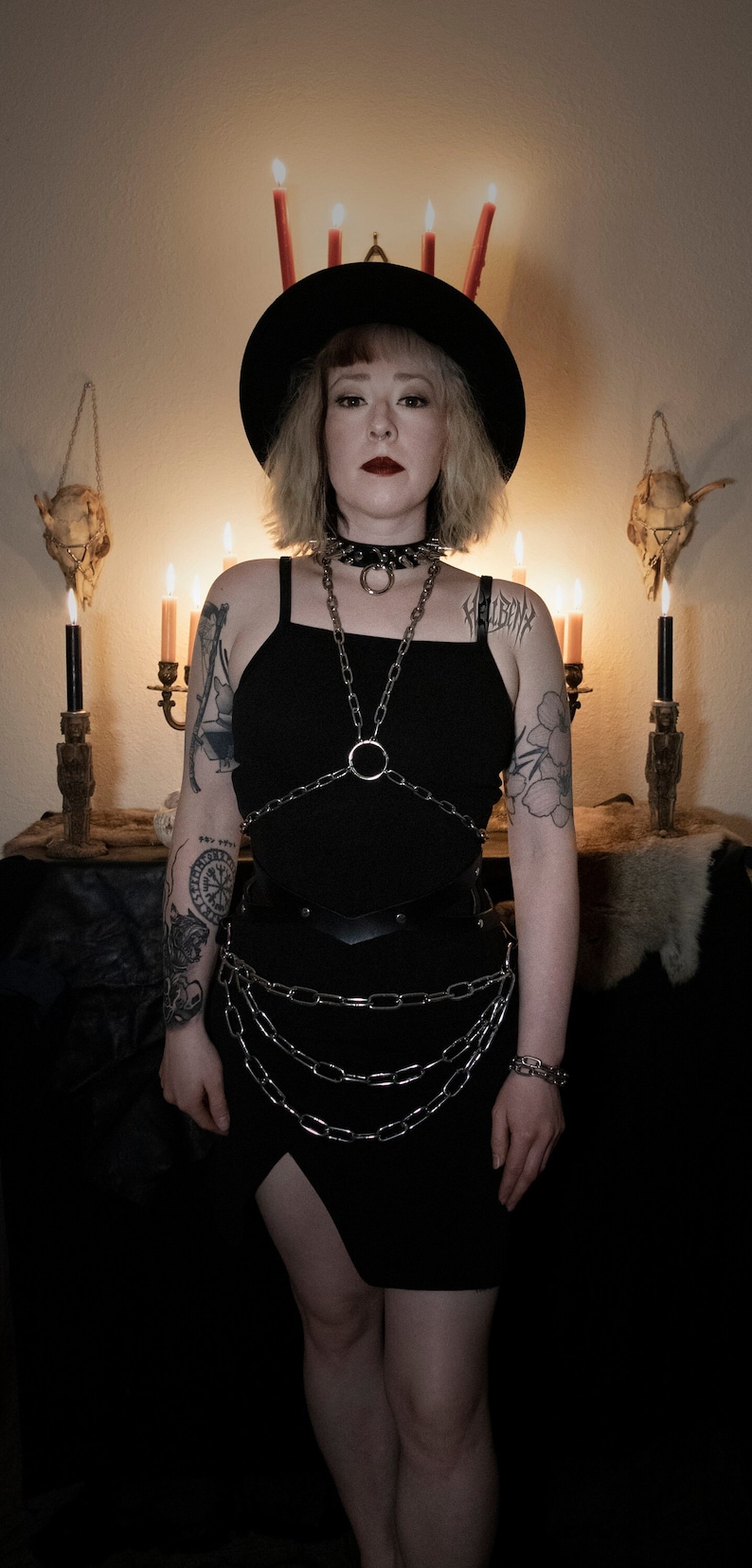 May include: A woman wearing a black dress with silver chains and a black hat. She has a tattoo on her arm and is standing in front of a dark background with candles and skulls.